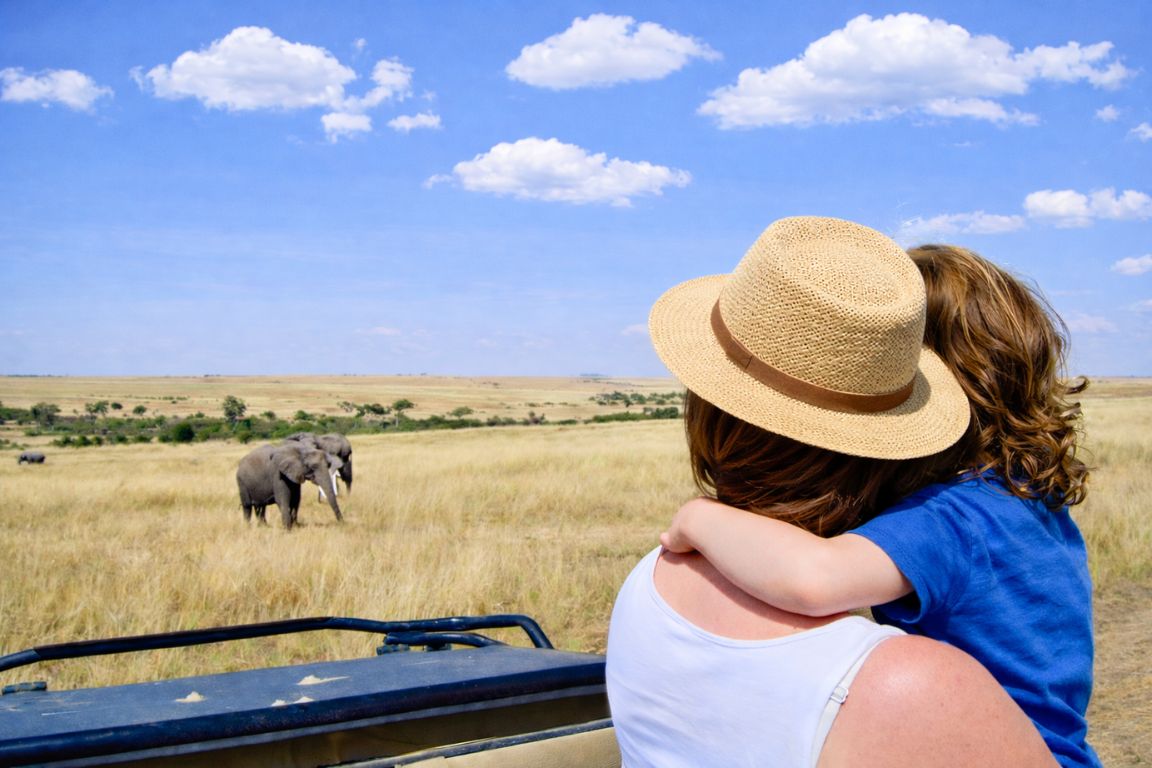 An adult holding a child in a safari vehicle watching an elephant on the Maasai Mara plains